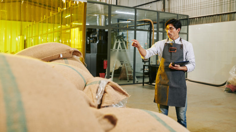 Roastery owner with digital tablet in hands counting sacks with coffee beans in warehouse